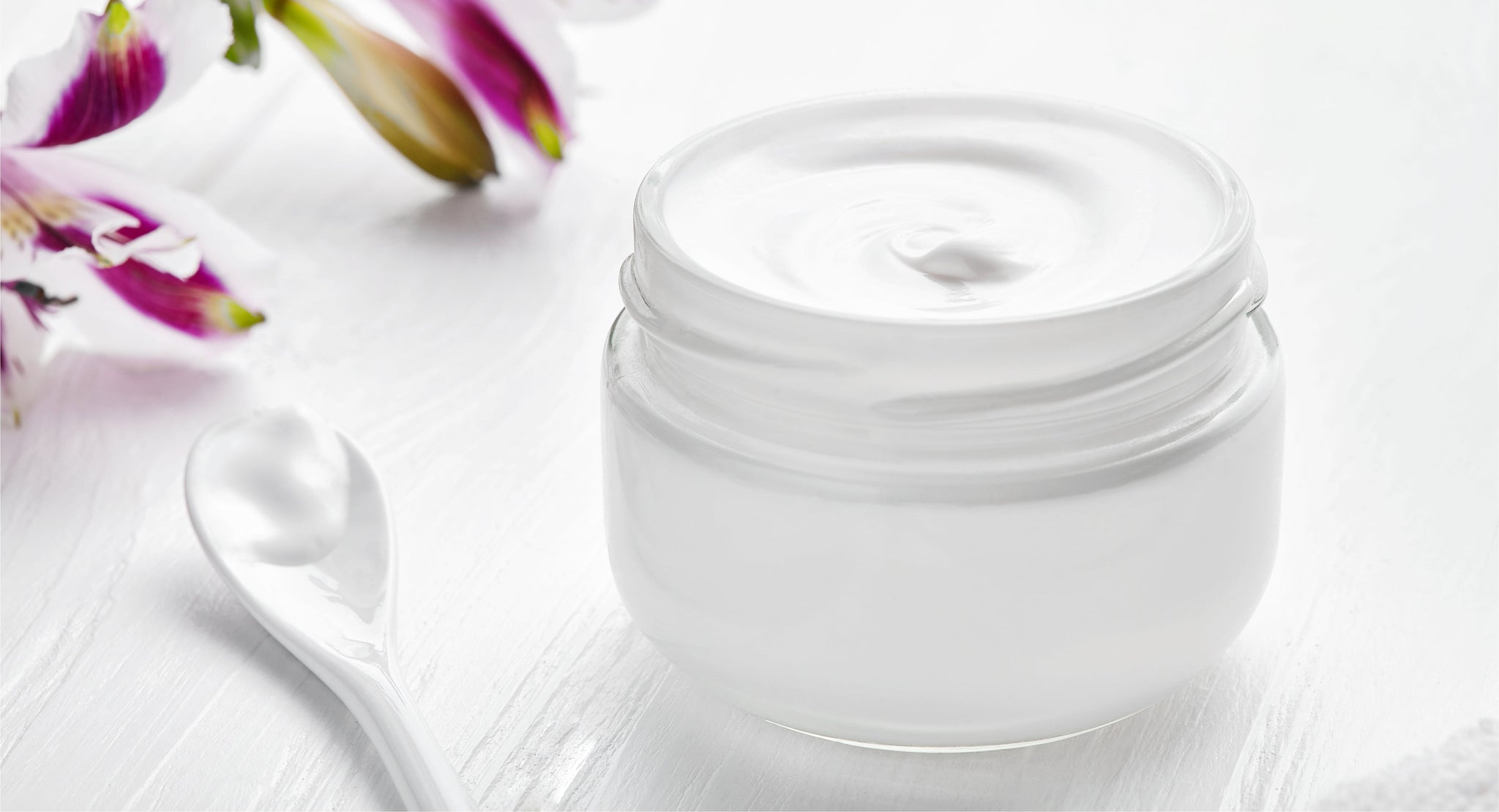 An open jar of white cream beside purple flowers.