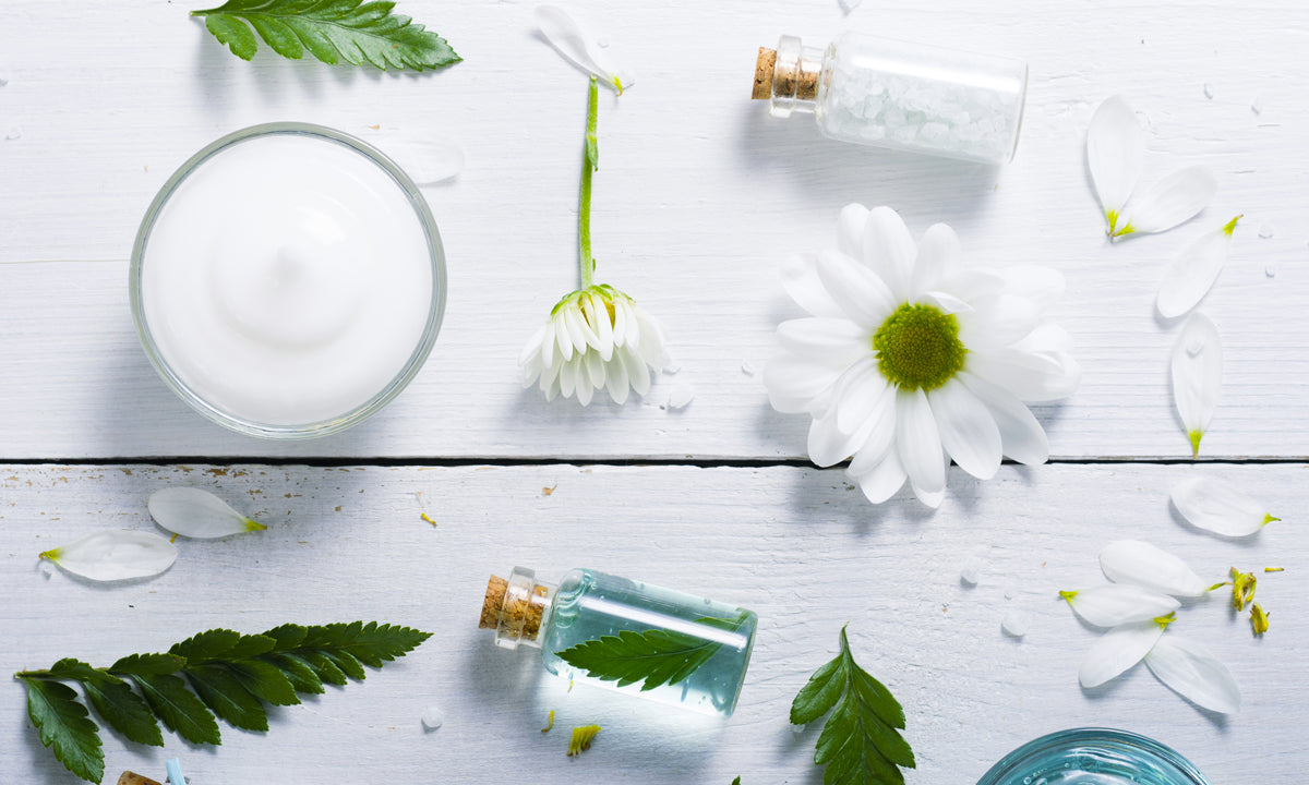 Bowl of moisturizer with white flowers and glass skincare bottles on a white surface