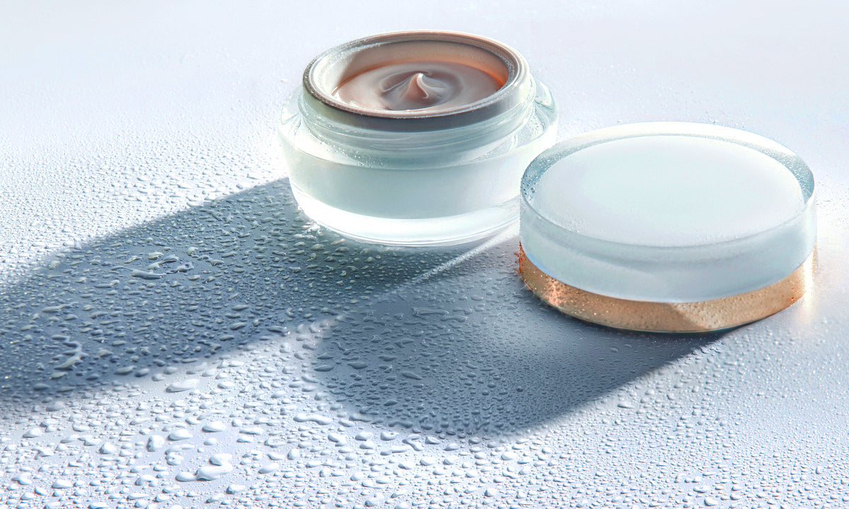 An open white moisturizer jar and lid on a surface with water droplets.