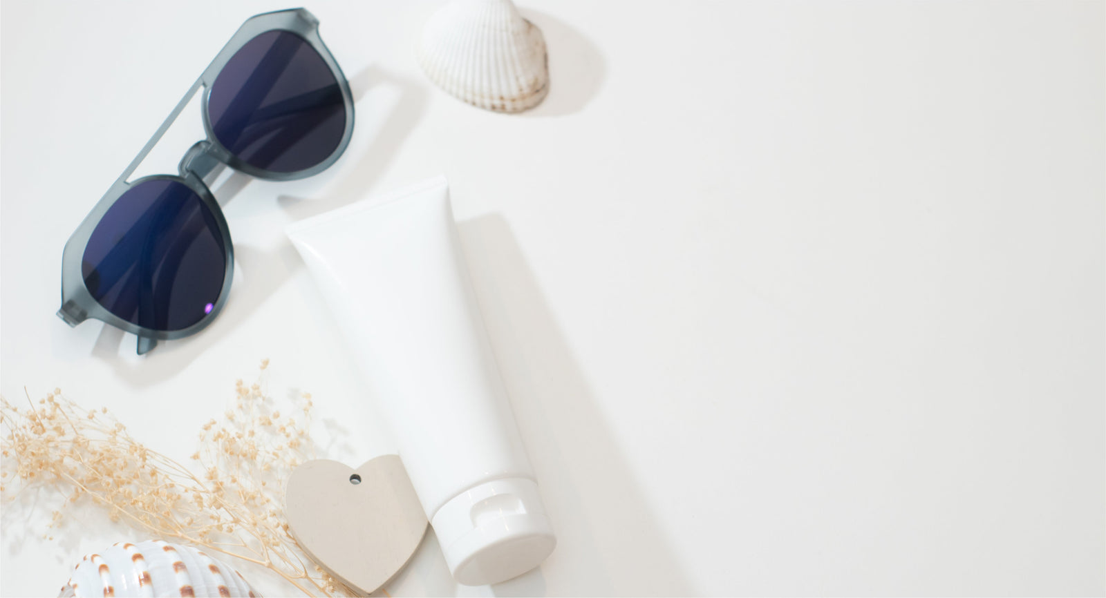 Unlabeled skincare tube beside sunglasses and seashells on a light surface